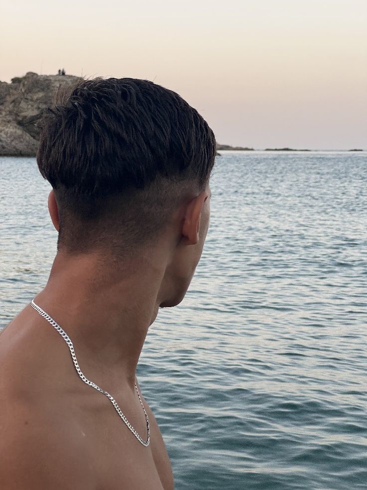 Homme torse nu portant une chaîne en argent, regardant la mer au coucher du soleil.