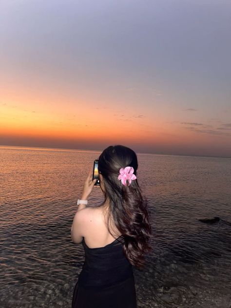 Femme de dos avec fleur rose dans les cheveux, photographiant un coucher de soleil sur la plage